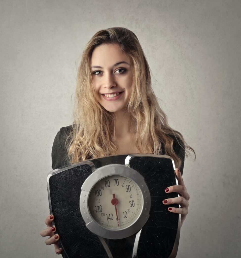 pexels-photo-3775540-3775540 Young woman with blond hair smiling, holding a vintage bathroom scale indoors.